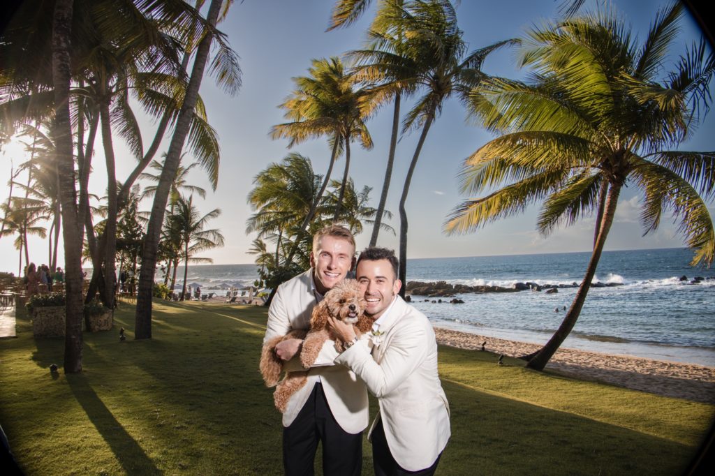 Two men in white suits, smiling and holding their beloved pet, a brown dog, stand on a lawn with palm trees near a beach under a clear blue sky.