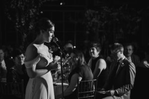 A bride is speaking into a microphone at a wedding held at the Wallace and Loeb Boathouse.