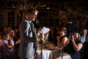 A man speaking into a microphone at a wedding reception by the Wallace and the Loeb Boathouse.