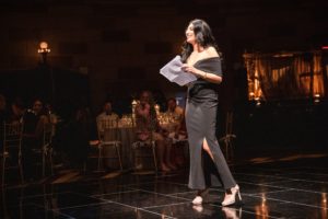 A woman in a black dress standing on the Gotham Hall stage.