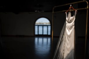 A Brooklyn wedding dress hangs on a hanger in a dark room during the summer.