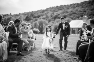 A little girl, dressed in a pretty summer dress, joyfully walks down the aisle at a Riverside Farm wedding.