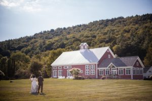 A summer wedding with the bride and groom standing in front of a red barn at Riverside Farm.