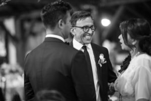 A man in a suit and tuxedo smiles at the bride and groom during their summer wedding at Riverside Farm.