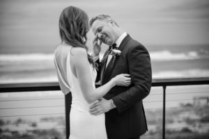 A newlywed couple, dressed in wedding attire, hugging on a balcony overlooking the ocean at their romantic New York wedding.
