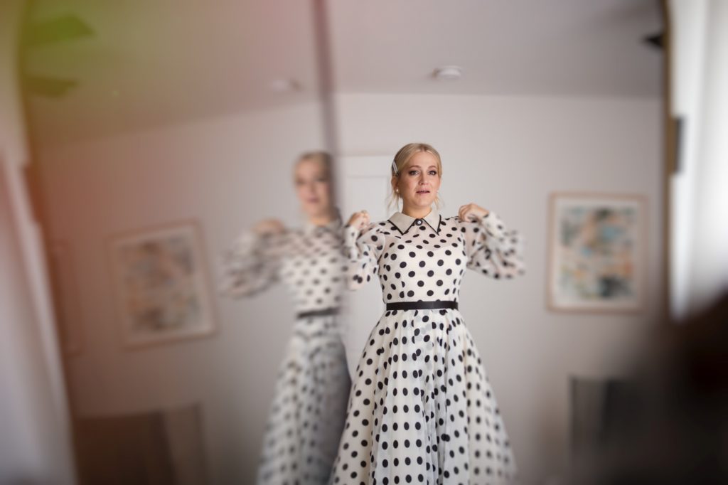 A woman in a polka dot dress is standing in front of a mirror, possibly preparing for a wedding in New York.