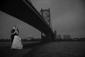 A bride and groom standing on a dock with a bridge in the background during their wedding in New York.