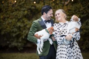A couple holding two pets, dressed in formal attire with smiling expressions, standing outdoors with greenery in the background.