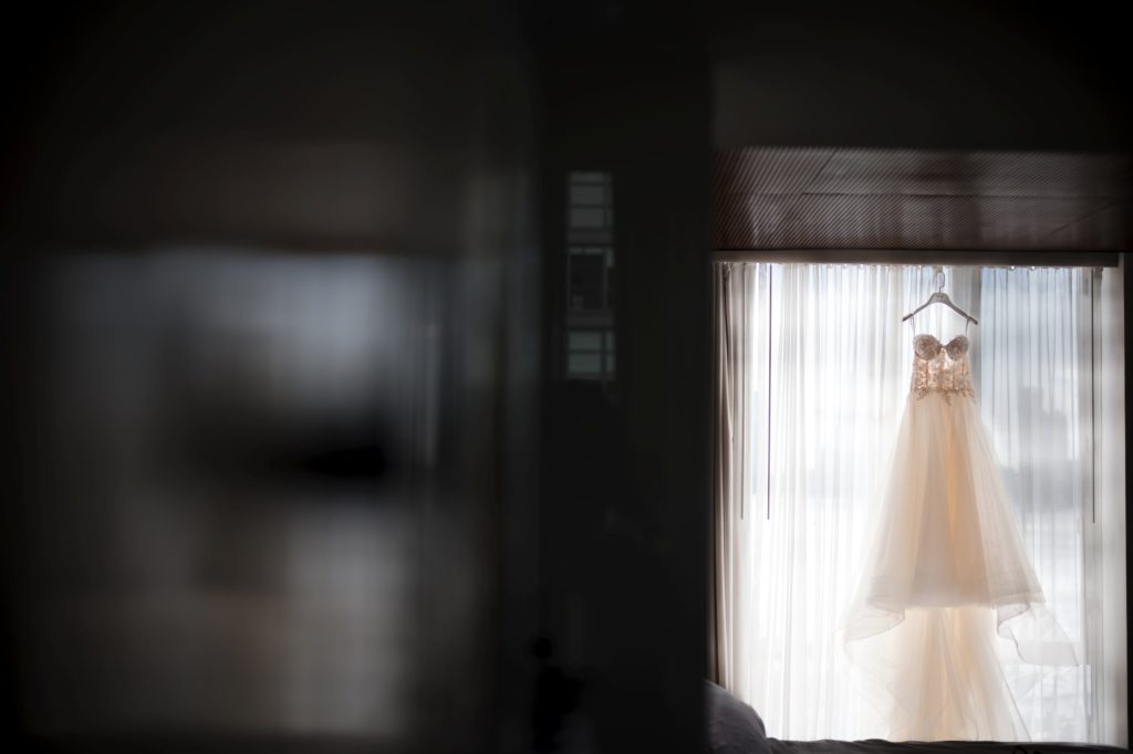 A new york wedding dress hangs from a window in a dark room.