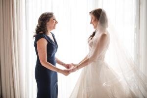 A bride and her mother, dressed in elegant wedding attire, holding hands in front of a picturesque window overlooking the beautiful cityscape of New York.