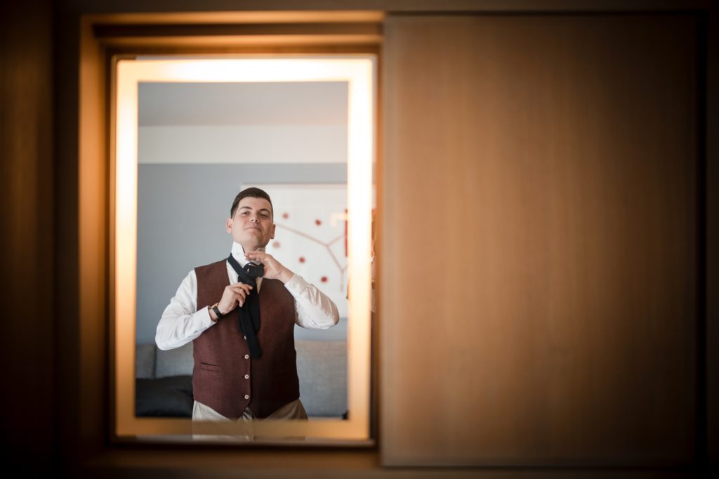 A man getting ready for his wedding in New York, meticulously putting on his tie in front of a mirror.