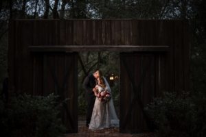A bride and groom standing in front of a wooden barn during their wedding at dusk in New York.