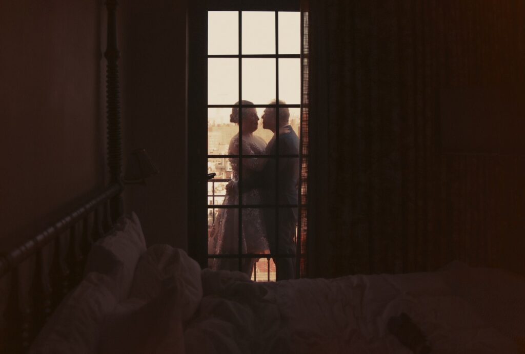 A newlywed couple standing in front of a window in a New York hotel room after their wedding.