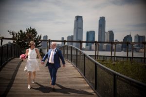 A newlywed couple walking on a boardwalk in front of the New York City skyline on their wedding day.