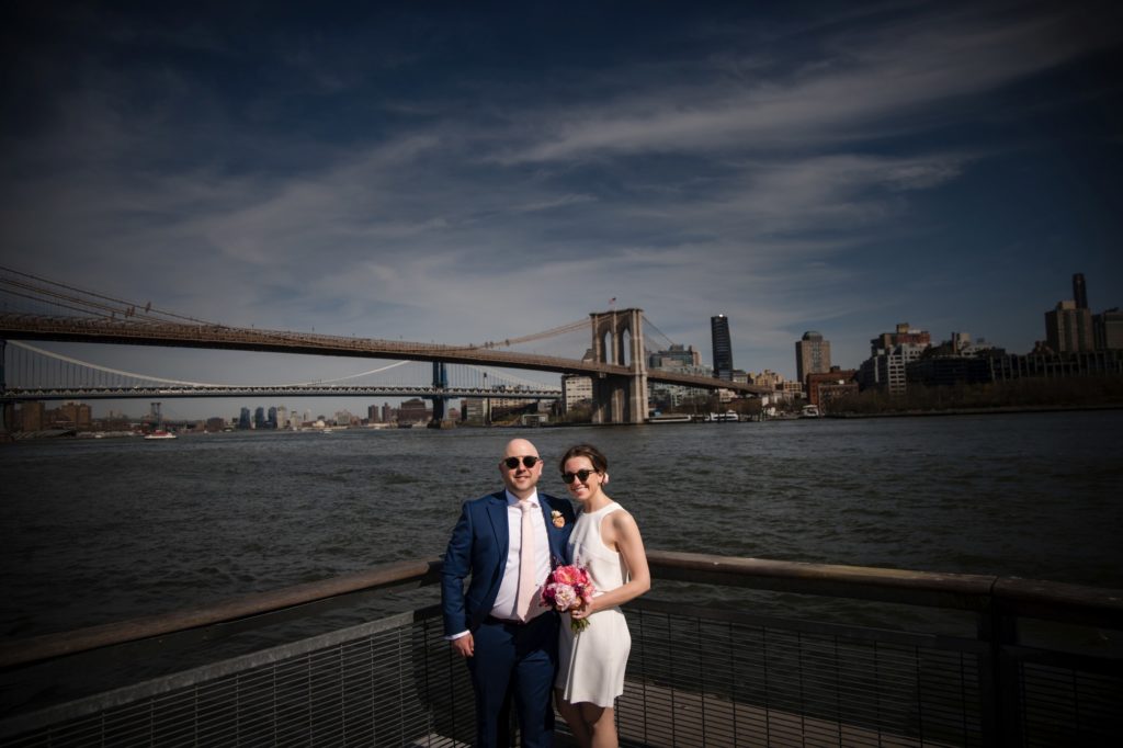 A bride and groom standing on a dock with the Brooklyn Bridge in the background, capturing their memorable wedding in New York.