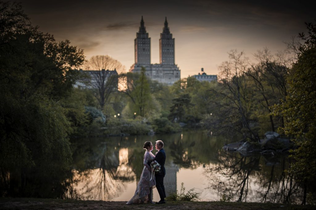 A newlywed couple standing in front of the picturesque lake in Central Park, New York, during a breathtaking sunset for their wedding ceremony.