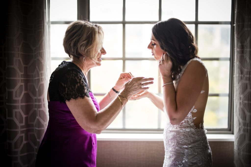 A bride and her mother, both dressed in elegant wedding attire, share a tender moment as they gaze at each other through a window.