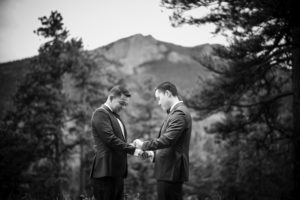 Black and white wedding photo of two men holding hands in front of a mountain.