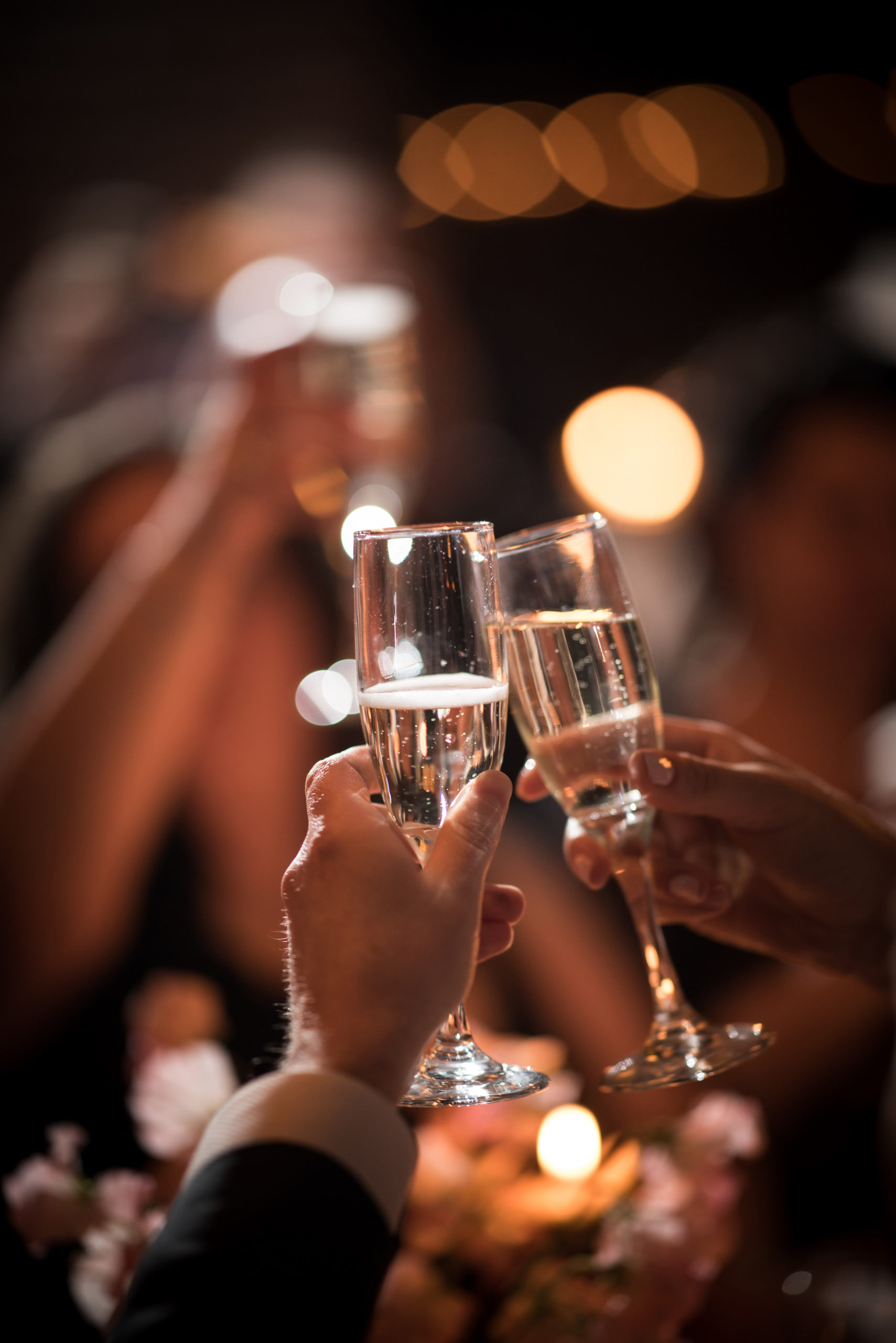 Close-up of hands holding and clinking champagne glasses in a celebratory toast, with blurred people and warm lights in the background.