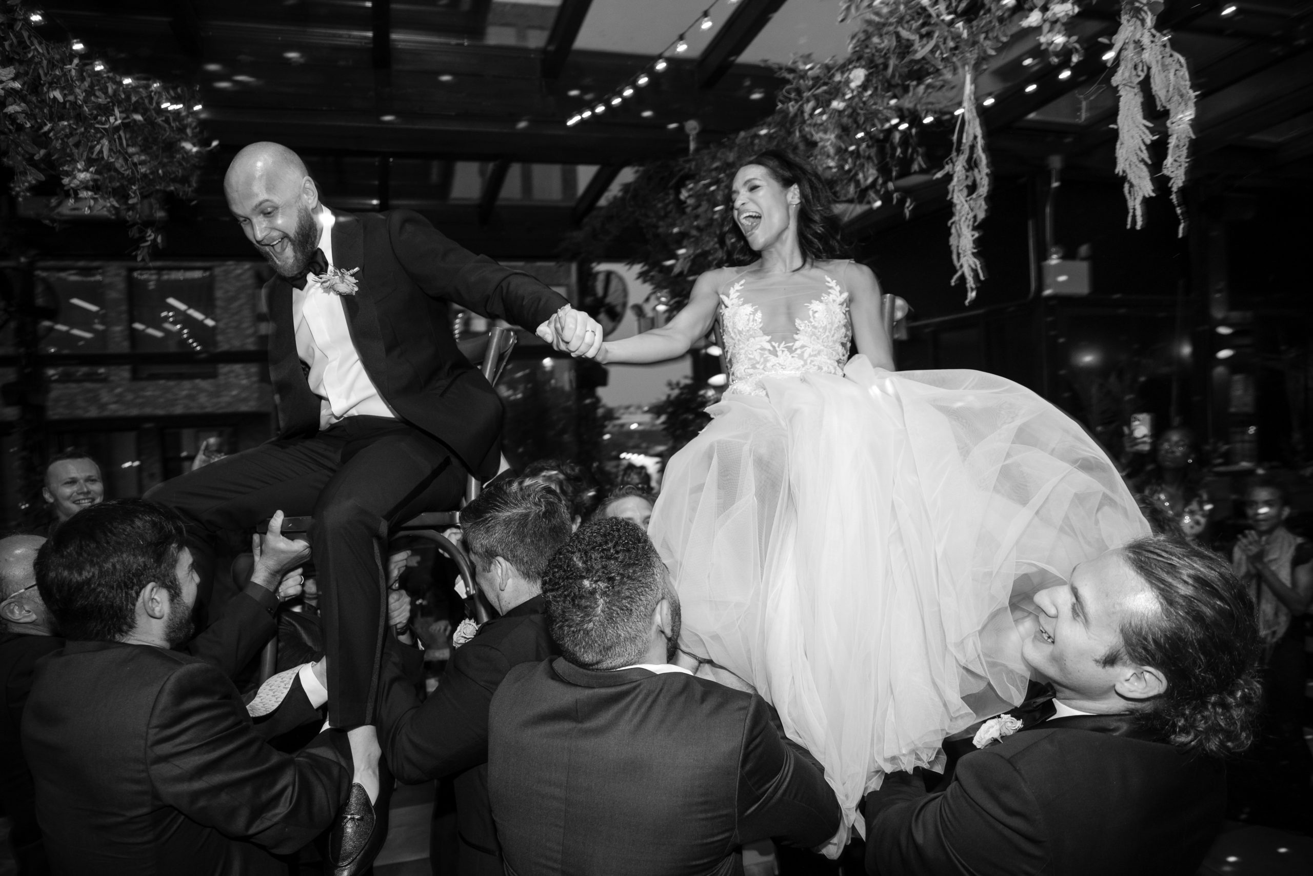 A bride and groom are lifted on chairs by guests during a wedding celebration inside a decorated venue.