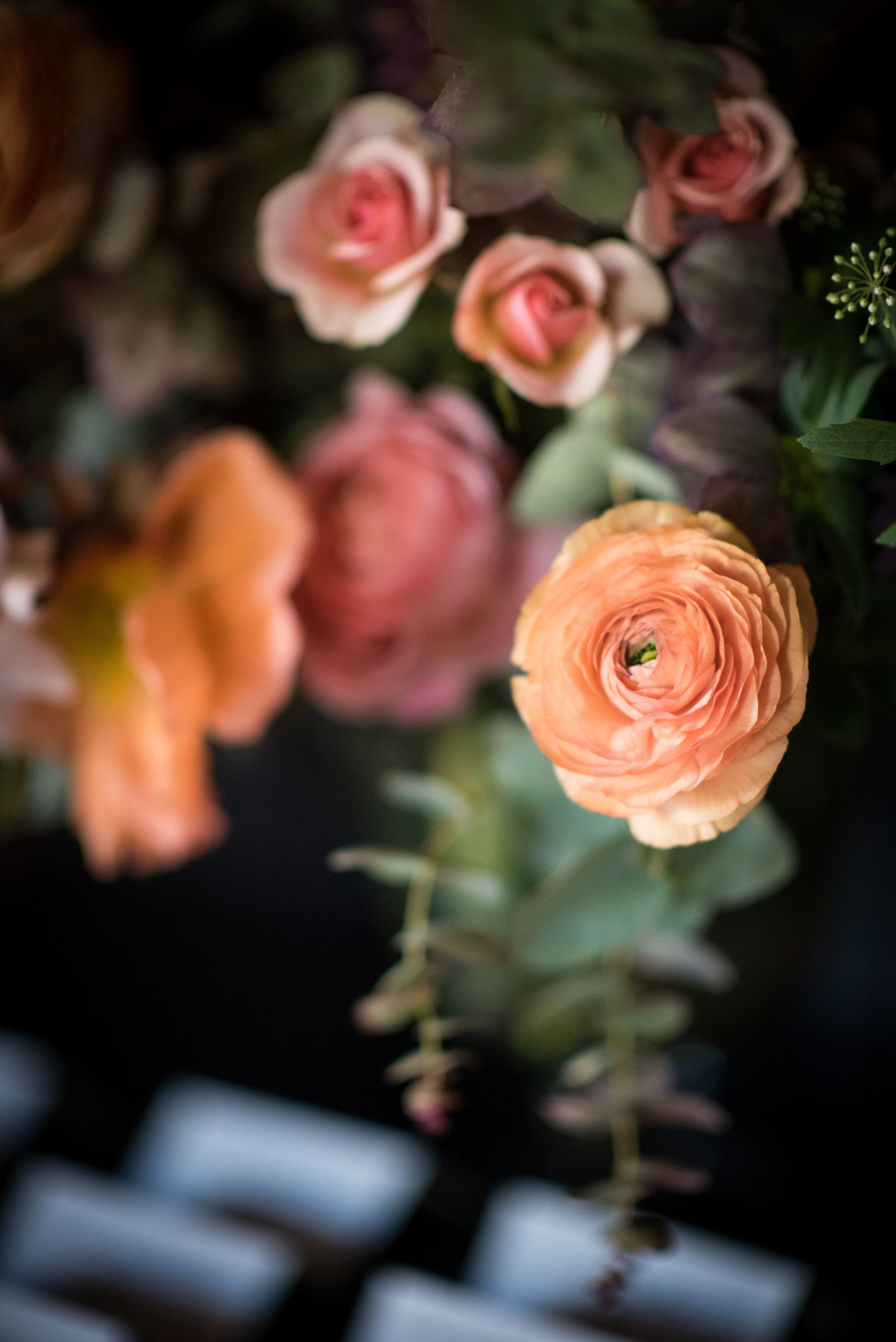 Close-up of assorted peach and pink ranunculus and rose flowers with greenery, softly focused in the background.