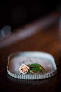 A single pale pink rose with green leaves rests on a rectangular, wavy-edged metal tray placed on a wooden surface.