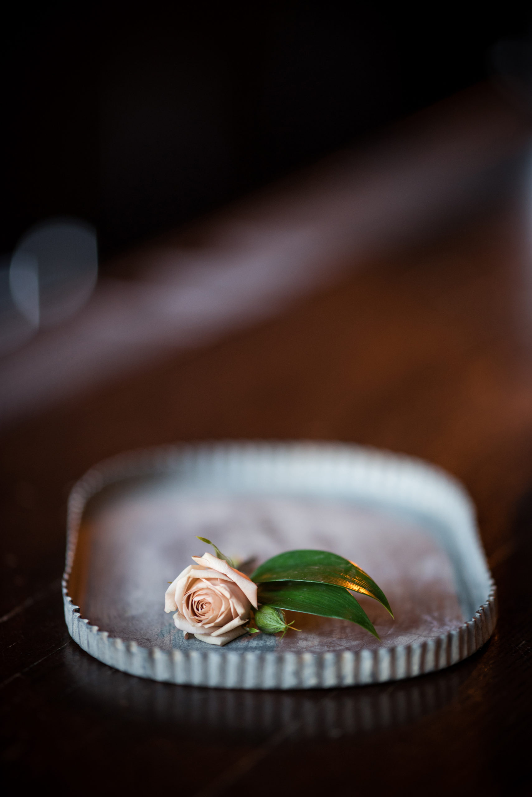 A single pale pink rose with green leaves rests on a rectangular, wavy-edged metal tray placed on a wooden surface.