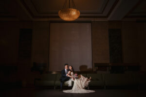 A bride and groom sitting on a couch together as she leans back into him.