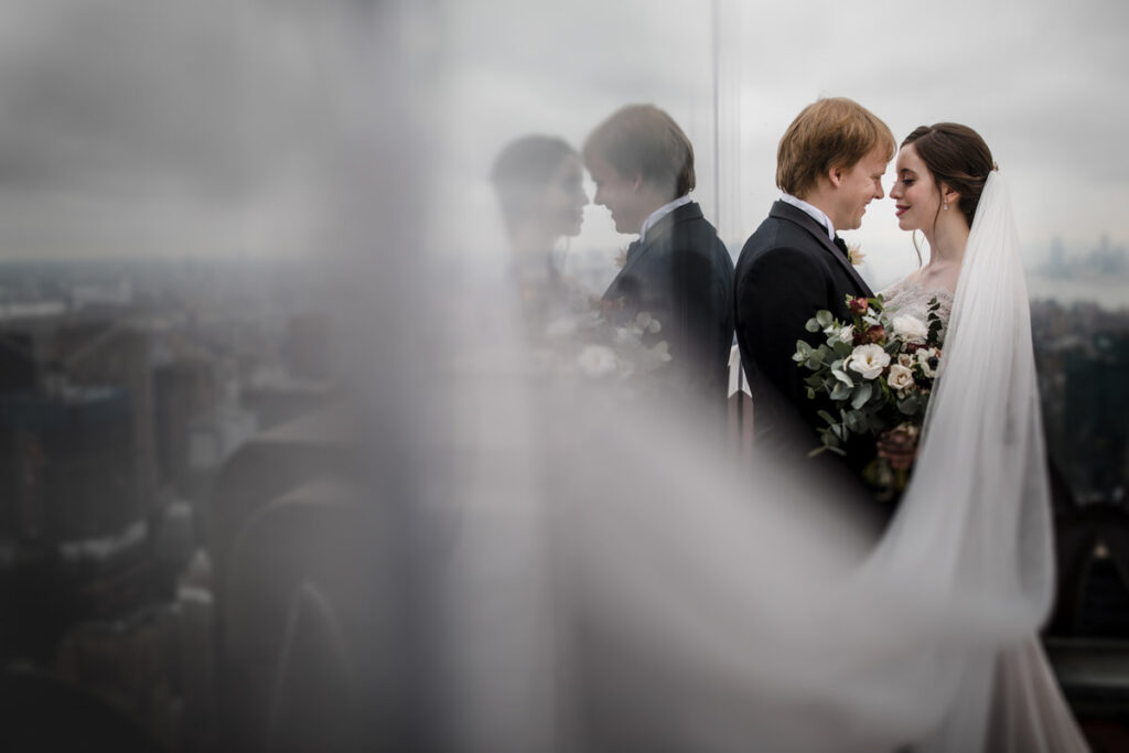 A groom leaning up against the side of a building as his bride leans in for a kiss.