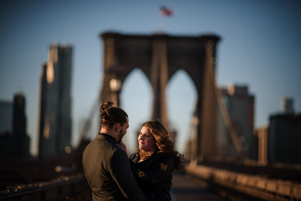 A couple with their arms around each other as they stand on a city bridge.