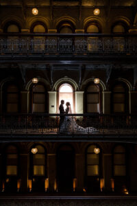 A bride and groom standing on a balcony with light shining through a door behind them.