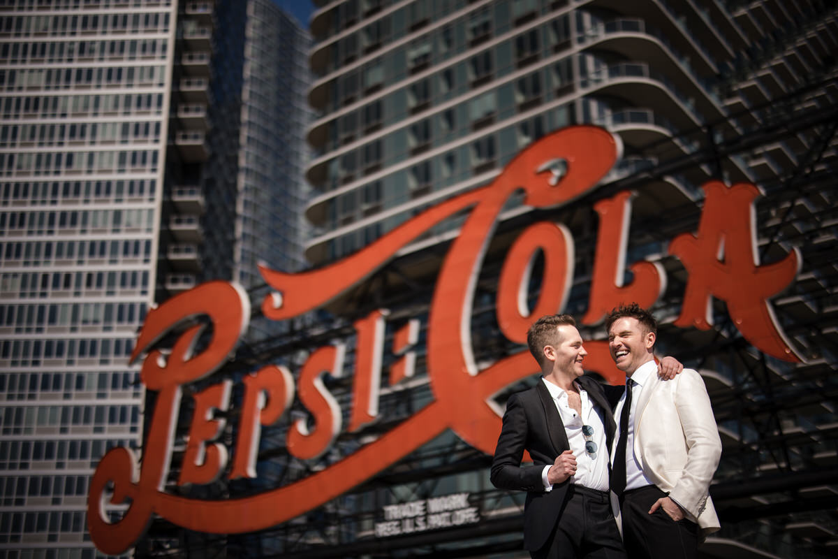 Two grooms with their arms around each other smiling in front of a large, red Pepsi-Cola sign. 