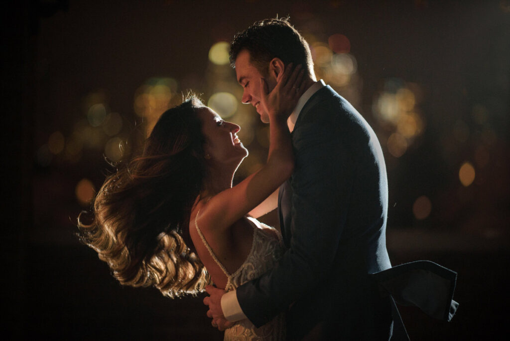 A bride and groom embrace in front of the skyline on a balcony of the Bowery Hotel at night.