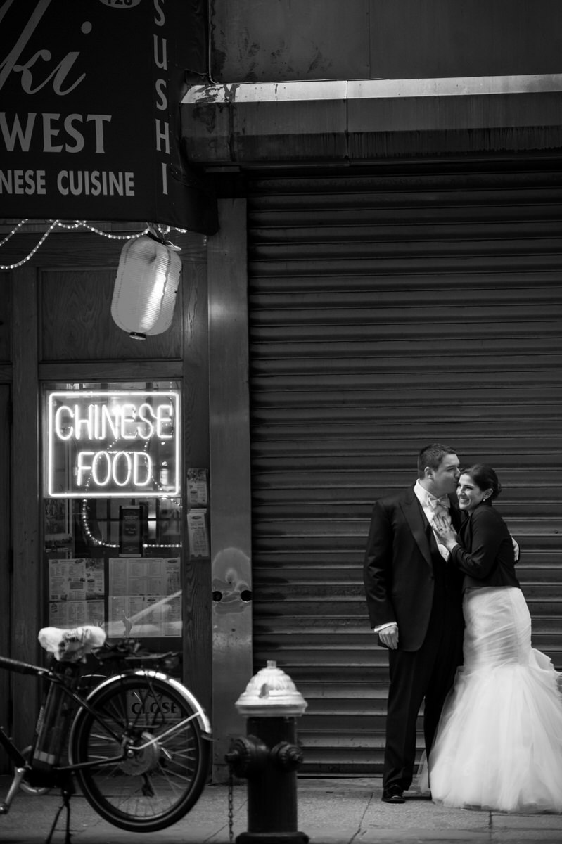 A groom kissing a bride on the cheek as they stand on a city street. 