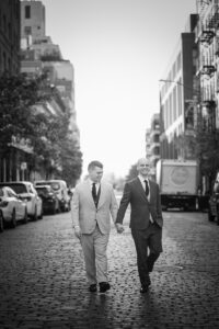 Two grooms holding hands and walking down a cobblestone city street.