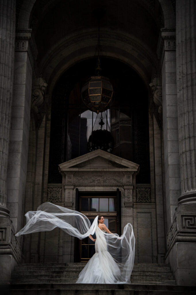 A bride standing at the steps of a large building while her veil flows in the wind.