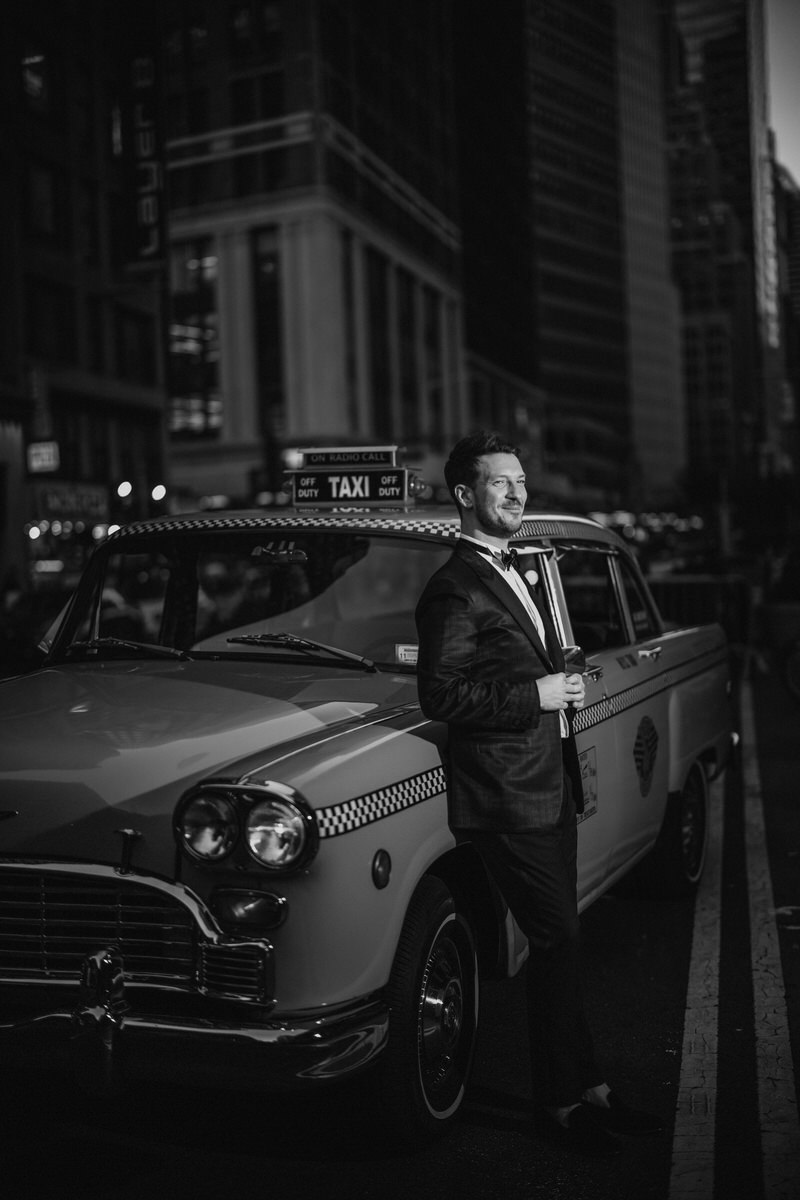 A groom in a tux leaning up against a vintage taxi. 