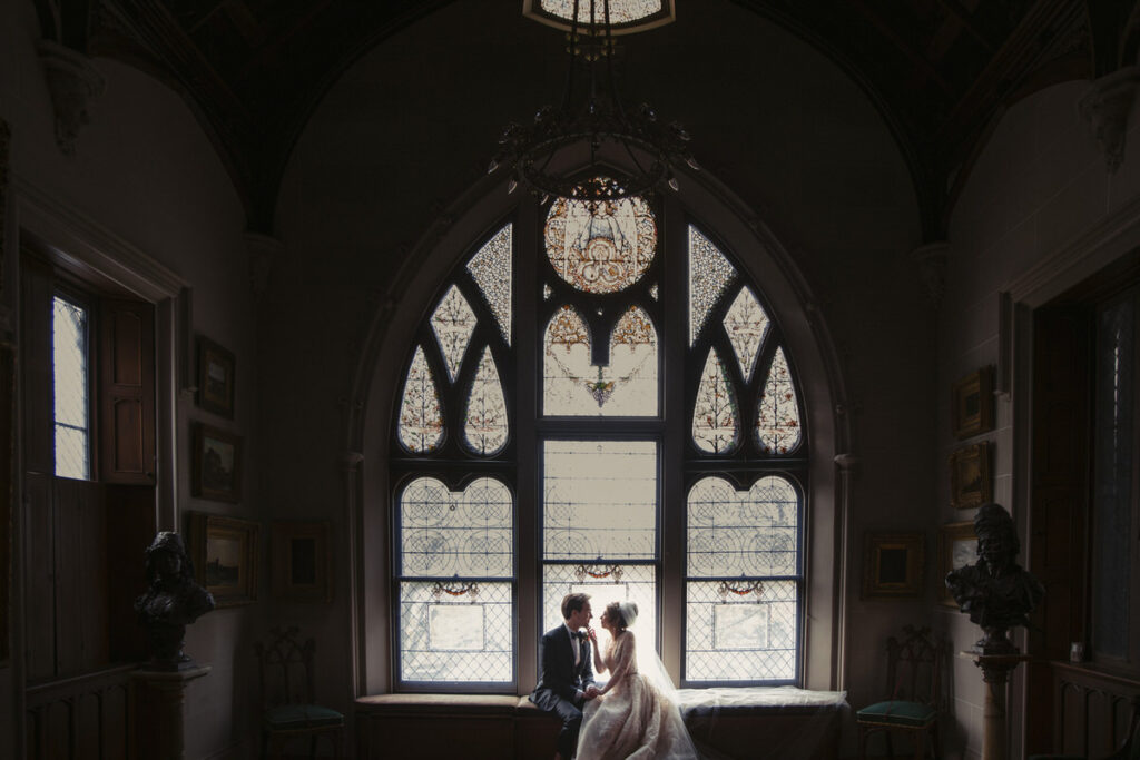 A bride and groom sitting in the windowsill of a large arched window.
