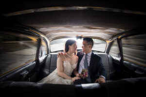 Newlyweds sharing an intimate moment in the backseat of a vintage car, with the bride in a lace gown and the groom in a gray suit. The blurred cityscape through the window suggests motion