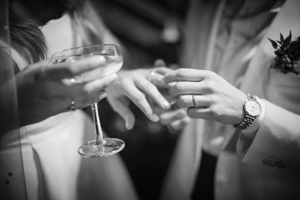 A close-up, monochromatic shot of a newlywed couple exchanging rings, with a focus on their hands and the wedding bands, set against the soft backdrop of their elegant attire.