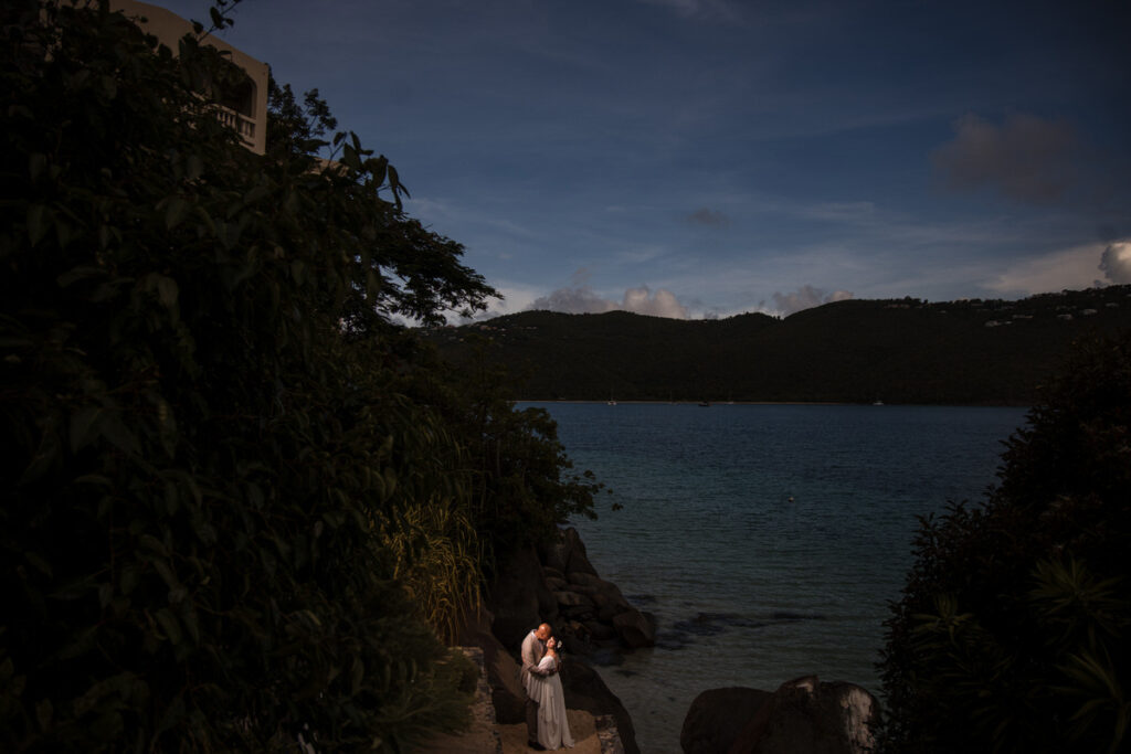 A couple embraces tenderly on a secluded rocky shore, with a serene bay and lush hills in the background, highlighting a private moment during their tropical destination wedding