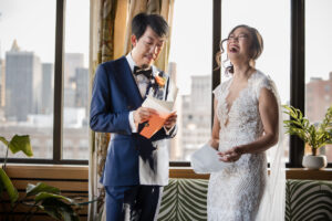 A bride and groom laugh heartily in a well-lit room with urban views, as the groom reads from an orange envelope, indicating a joyful, candid moment during their wedding day