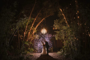 A couple stands beneath a fairytale-like chandelier in an enchanted forest setting, surrounded by twinkling lights and natural beauty, creating a magical wedding night atmosphere