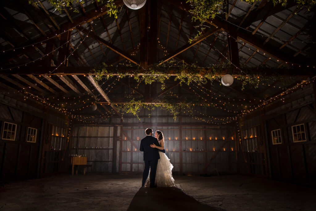 The newlyweds share a romantic dance in a rustic barn adorned with string lights and hanging greenery, their silhouettes illuminated by the soft ambient lighting, evoking a cozy and intimate reception