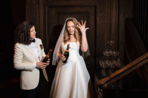Two brides celebrate their wedding, one in a tuxedo and the other in a white dress, playfully opening a bottle of champagne by a wooden staircase, with joy and anticipation in the air