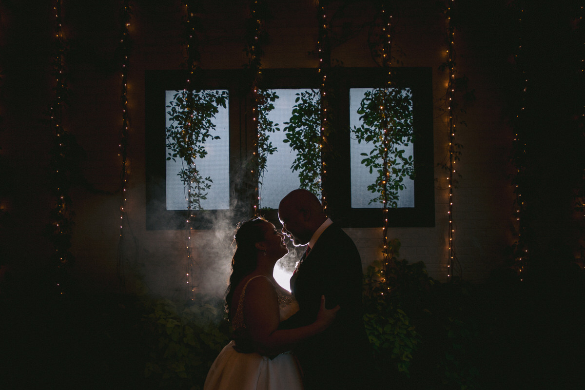 A silhouetted bride and groom embracing in front of bright windows, framed by ivy and twinkling lights, in a dimly lit romantic setting.