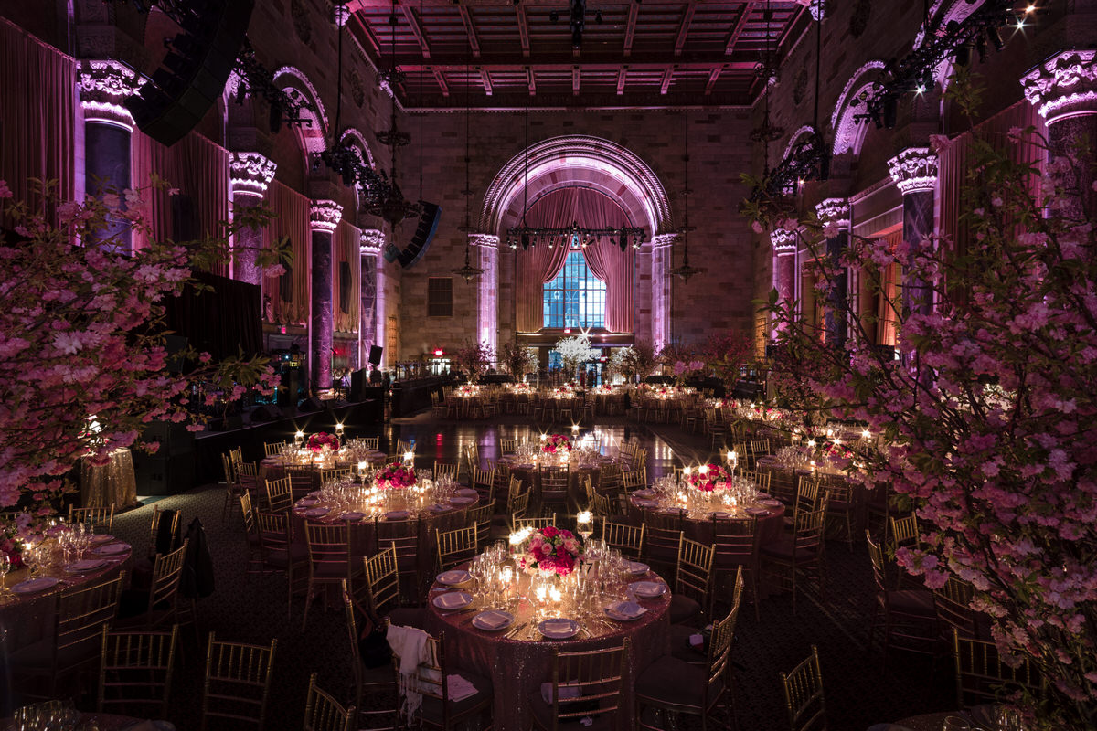 Opulent wedding reception hall with arched windows, grand chandeliers, and tables adorned with pink floral centerpieces and golden chairs.