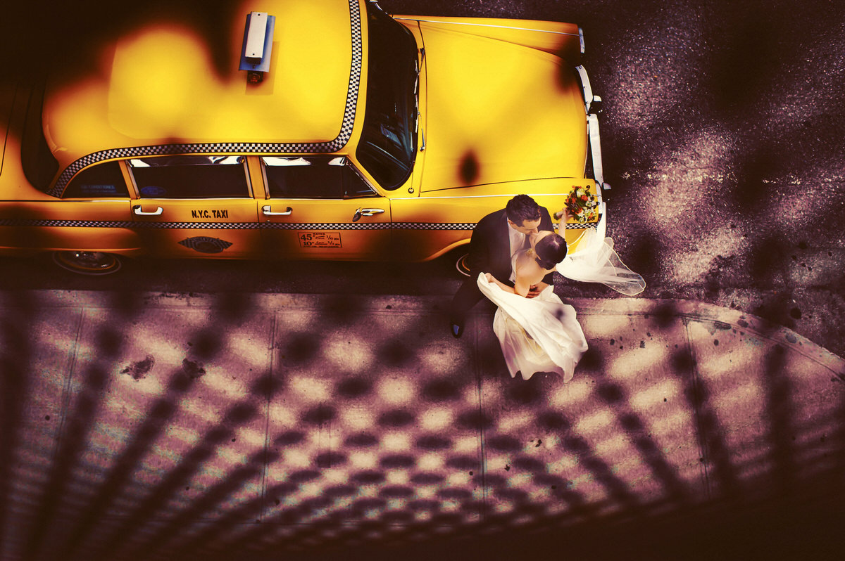 An overhead view of a bride and groom kissing on a New York City street, casting long shadows beside a yellow taxi cab.