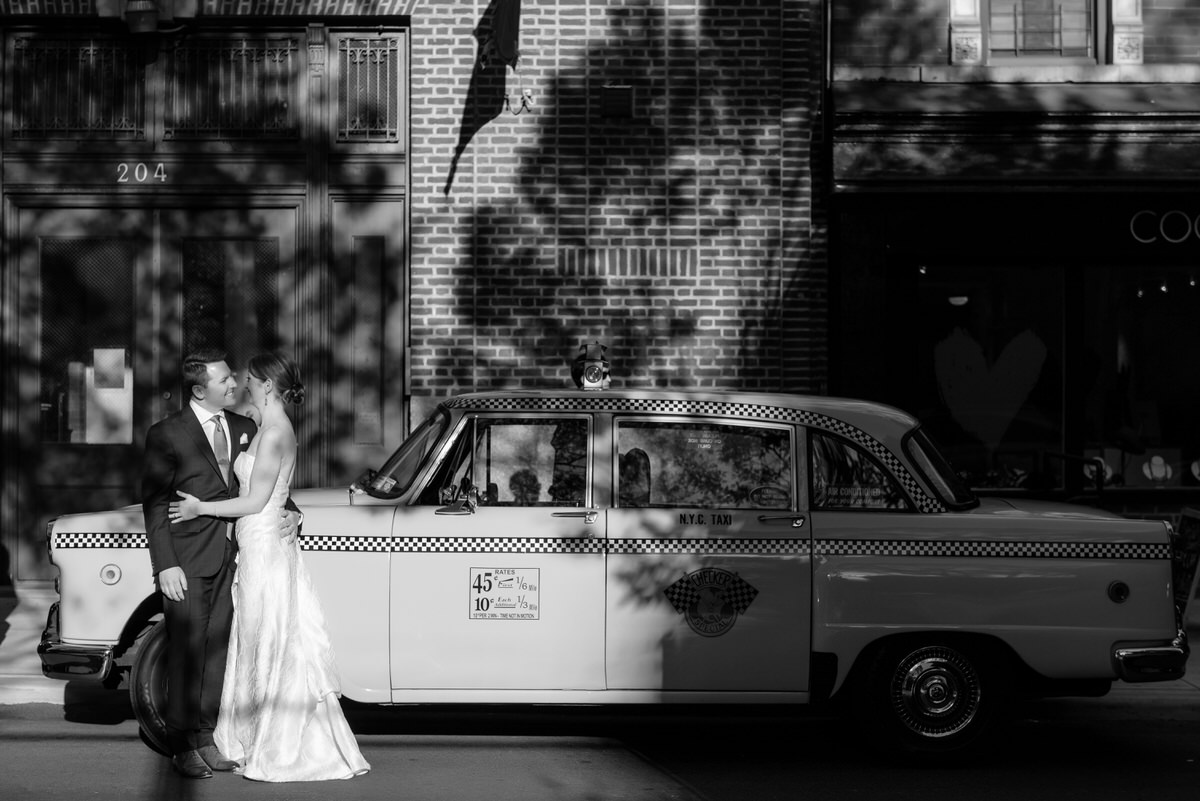 A classic black and white photograph of a newlywed couple sharing a moment in front of an iconic New York City checkered taxi.