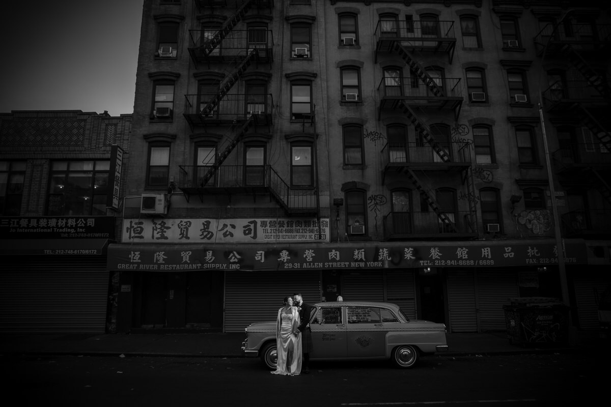 A monochrome shot of a bride and groom standing in a deserted city street, with a vintage taxi and old buildings, evoking a timeless New York vibe during their wedding at one of the luxury wedding venues in NYC